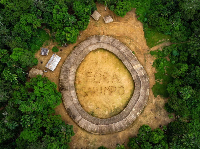 Vista aérea de uma maloca Yanomami. No centro, indígenas formam as palavras Fora Garimpo. Ao redor da maloca, existem muitas árvores de tom muito verde.